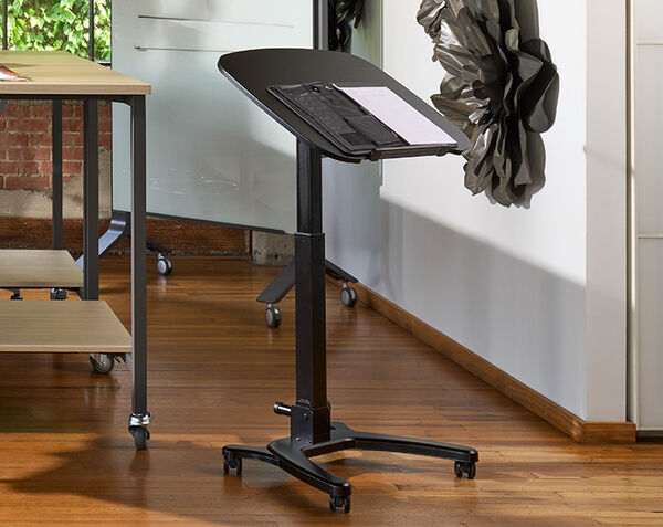 Mobile laptop desk with tabletop tilted at a steep angle holding a keyboard and papers with a binder clip, shown in an office space near a wood table and a whiteboard, illustrating the adjustable tilt feature.