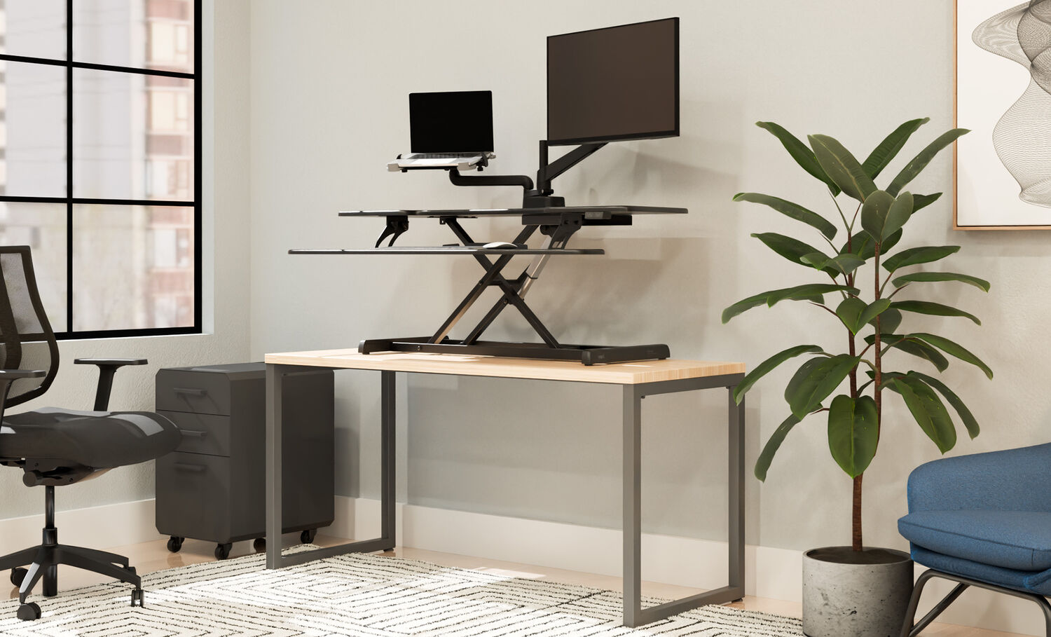 Black electric standing desk converter raised on a wood desk, holding a laptop and monitor, in an office with a mesh chair, filing cabinet, plant, and blue accent chair.