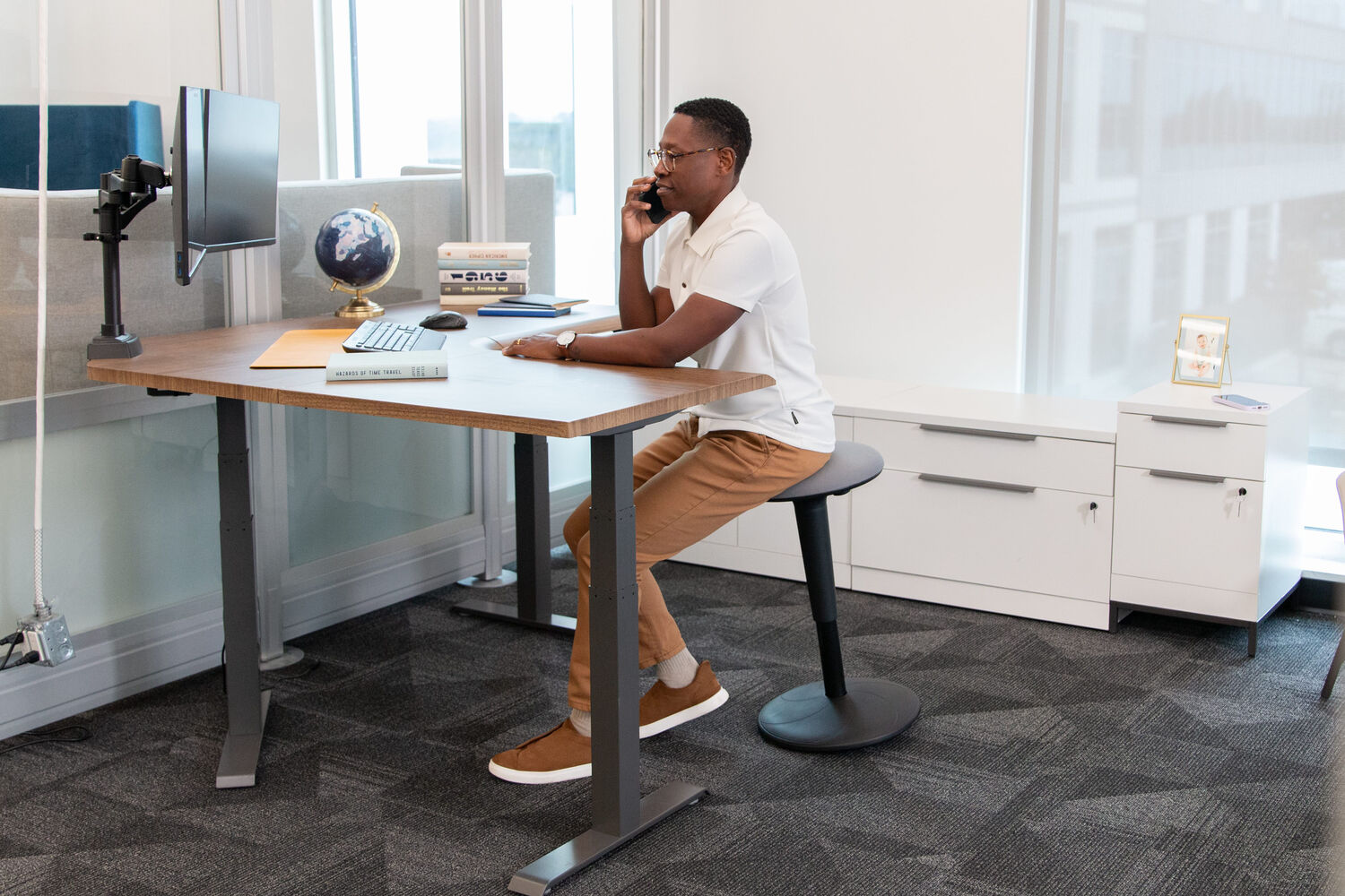 man sitting on a balance stool leaning forward while on the phone at an electric standing desk