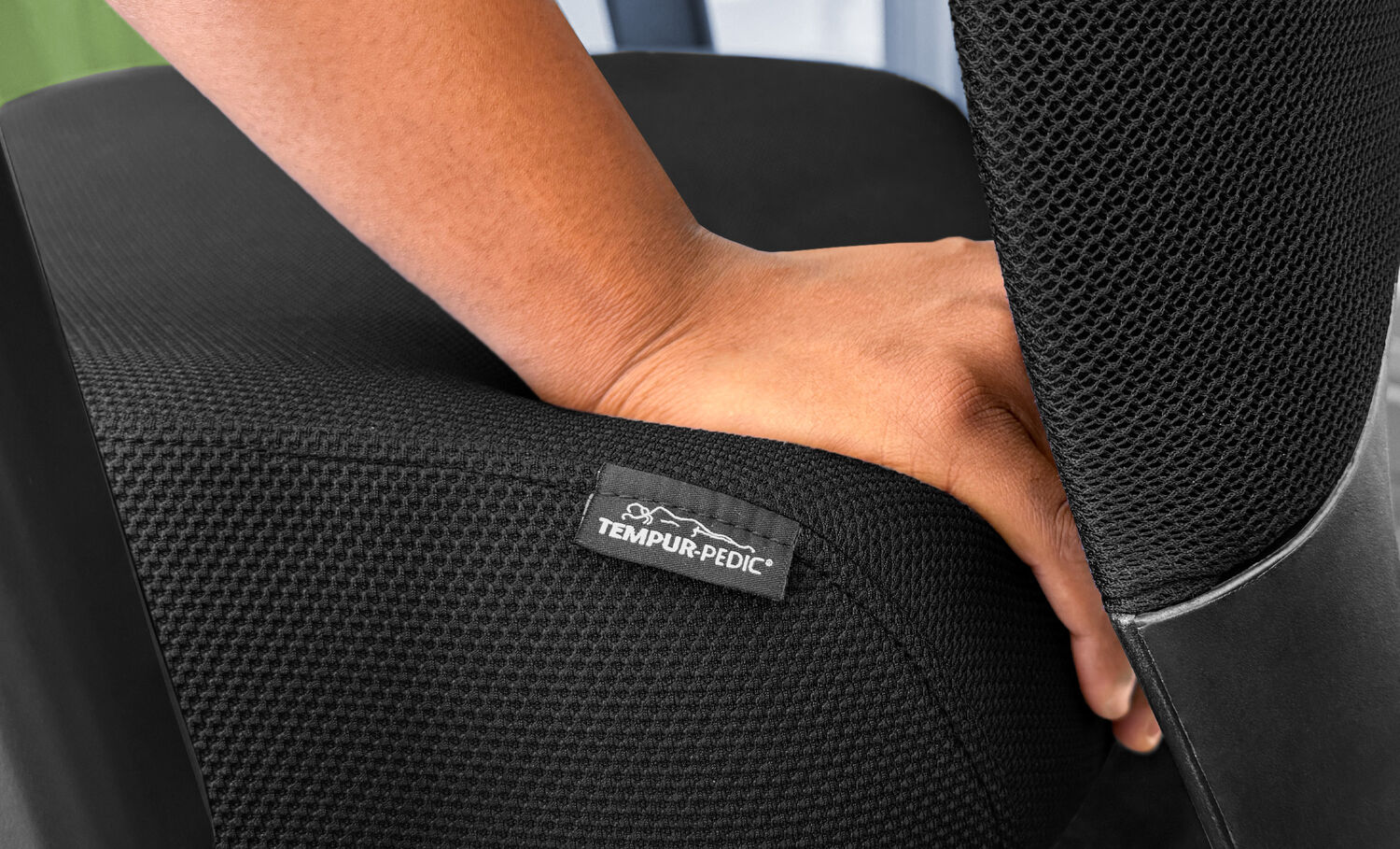 Close-up of a hand pressing into the black Vari TempurPedic seat cushion, showing the material's softness and support, with a branded fabric tag visible on the textured surface.