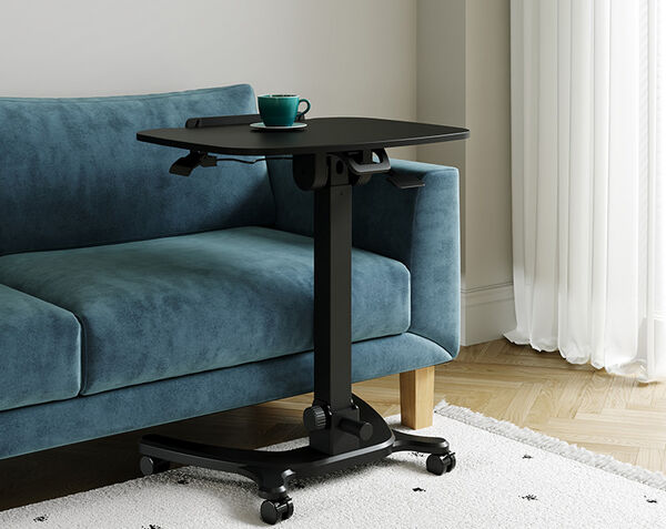 Mobile laptop desk with a teal mug and saucer on the tilted black tabletop, positioned beside a teal velvet sofa in front of a curtained window, showing the tilt lever and height-adjustment crank.