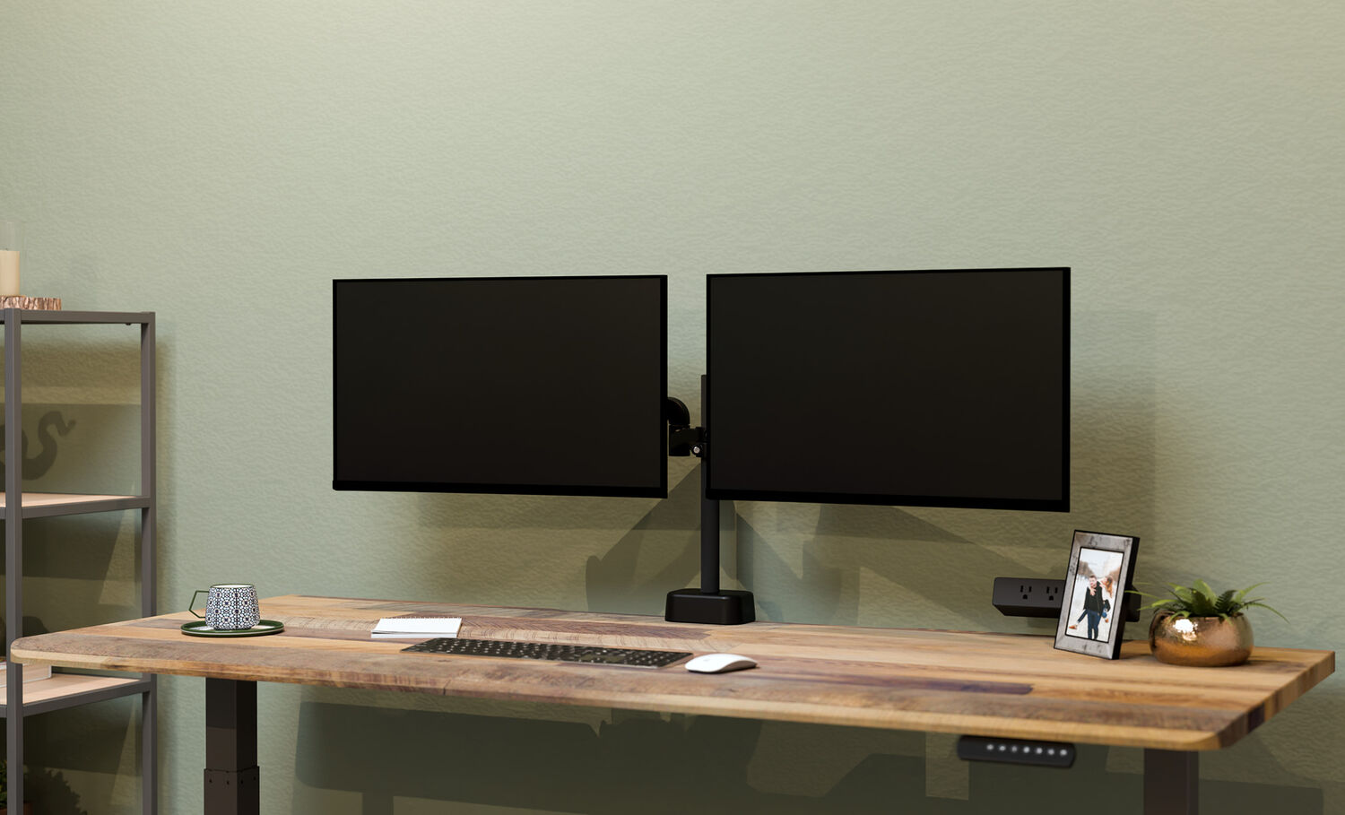 Two black monitors mounted side by side on a dual monitor arm against a sage green wall, above a wood desk with a keyboard, mouse, and picture frame.