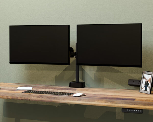 Two black monitors mounted side by side on a dual monitor arm against a sage green wall, above a wood desk with a keyboard, mouse, and outlet.
