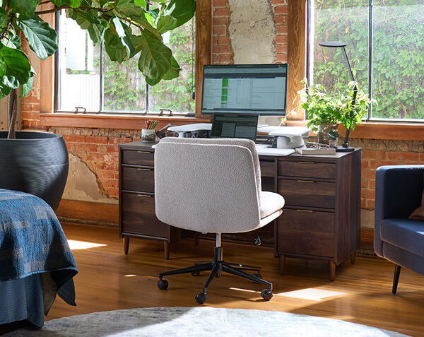 Back view of the light gray boucle armless office chair at a wood desk with a monitor and laptop, in a bright room with a fiddle leaf fig plant, a blue sofa, and large windows.