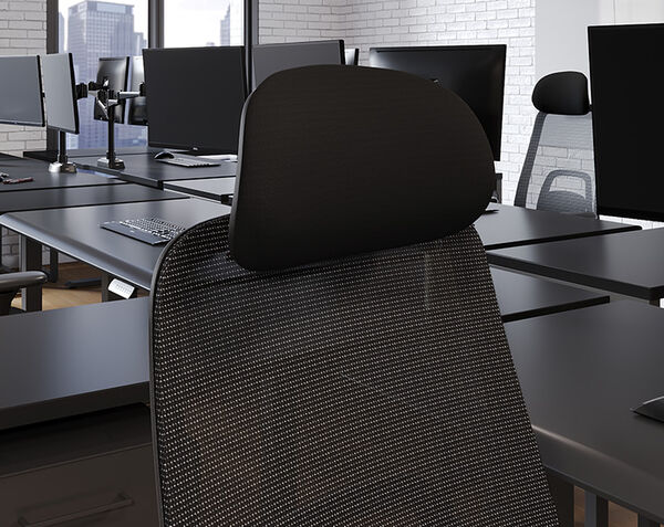 Close-up rear view of a black mesh office chair's headrest and upper backrest, shown at a desk in an open office with monitors in the background.
