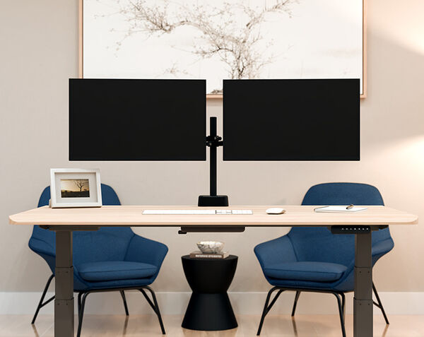 Two black monitors mounted on a dual monitor arm above a wood desk, flanked by blue accent chairs, with a framed tree print on the wall behind.