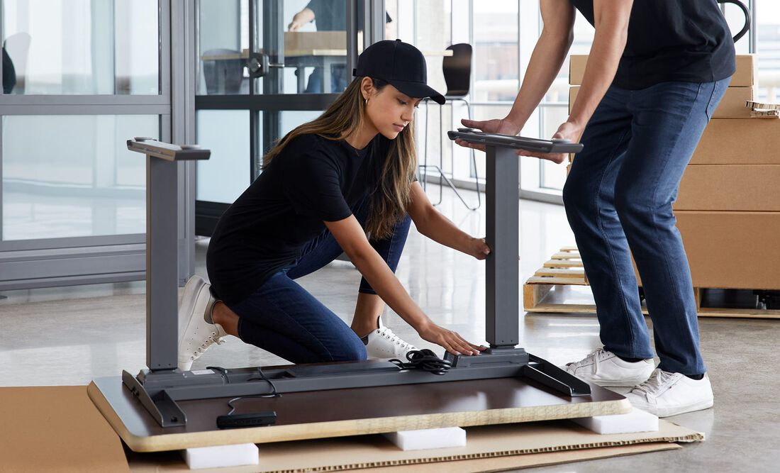 Two individuals attaching the legs to the Electric Standing Desk 60x30 during assembly in office setting