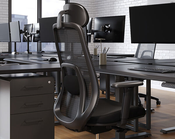 Rear angled view of a black mesh task chair with adjustable headrest, lumbar support, and armrests, positioned at a desk in an open office with monitors and a filing cabinet.