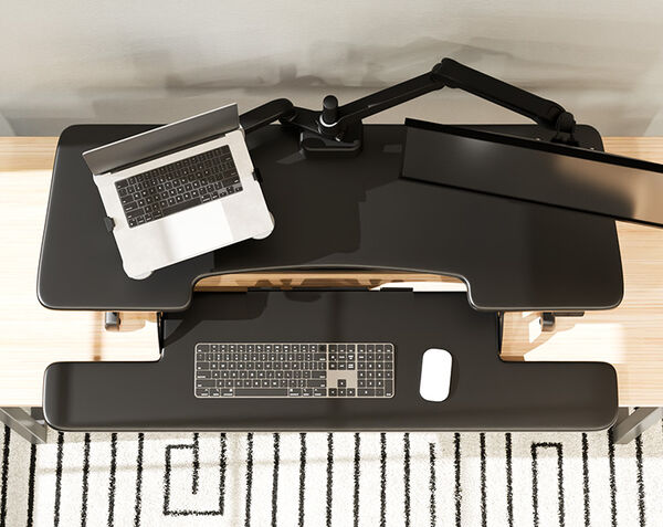 Top-down view of a black electric standing desk converter with a laptop tray, monitor arm, keyboard, and mouse, on a wood desk with a striped rug below.
