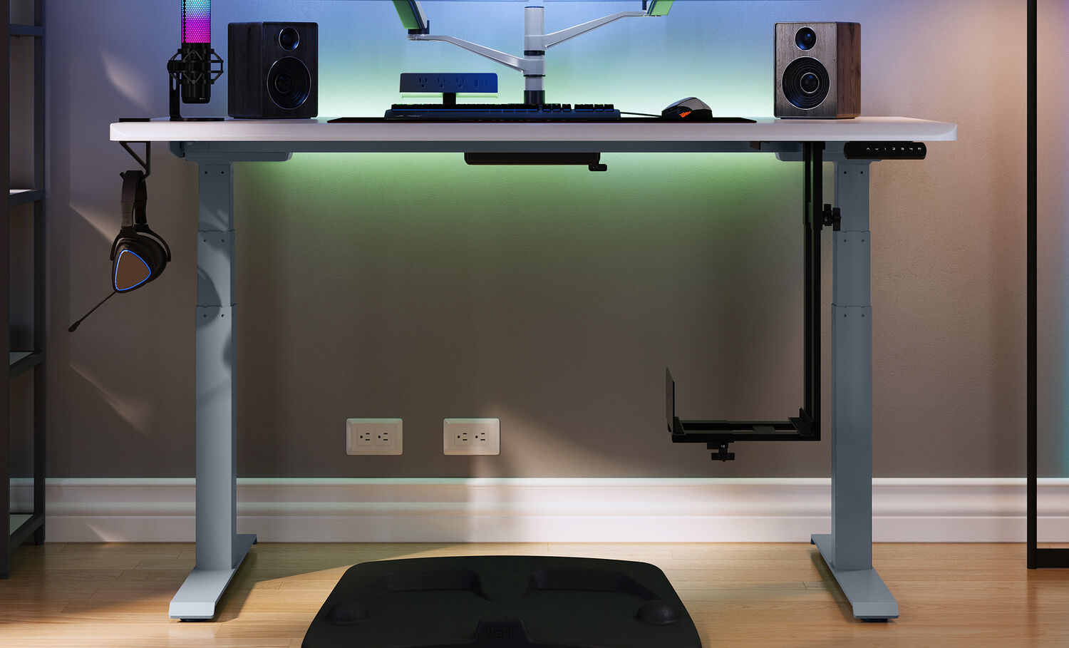 Modern gaming desk setup with RGB lighting, dual monitor arms, speakers, and a PC mount under the desk, plus a headset hanger and anti-fatigue mat on a wooden floor.