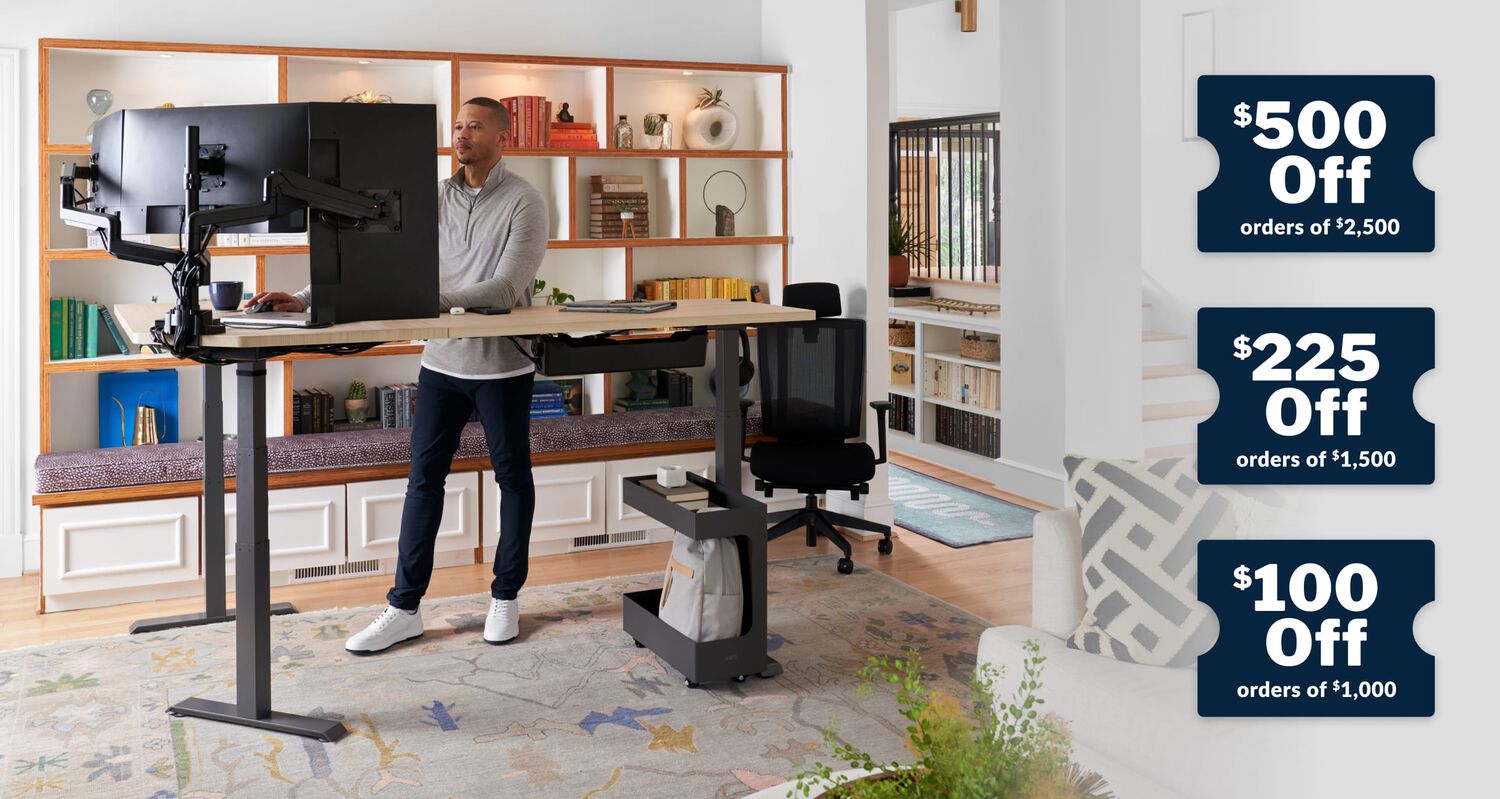 a man standing at an L shape electric standing desk and working in a home office and to the right of the picture tickets running vertically with five hundred dollars off of order of two thousand dollars two hundred and twenty five dollars off of order of one thousand dollars one hundred dollars off of orders of seven hundred and fifty dollars