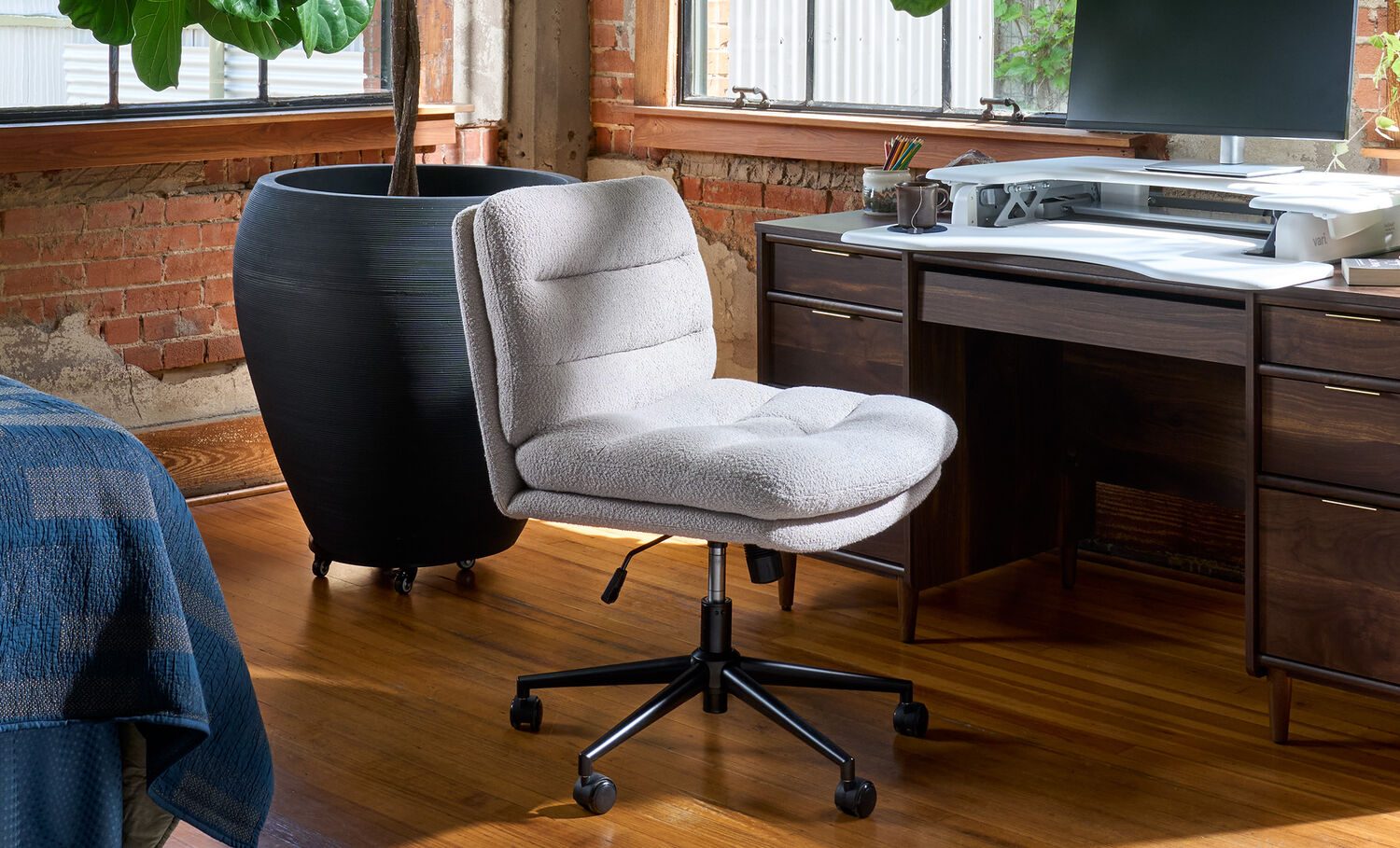 beige boucle armless office chair with a channel-tufted back and black five-star caster base, positioned in front of a dark wood desk with a monitor, laptop, and desktop converter, in a loft-style office with brick walls and large windows.