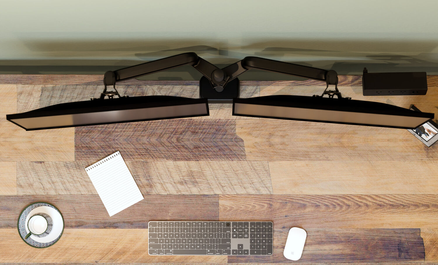 Top-down view of two black monitors mounted on a dual monitor arm, with a keyboard, mouse, notepad, teacup, and picture frame on a wood desk.