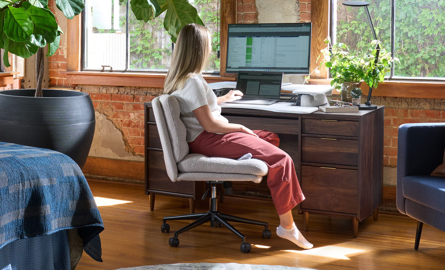 Woman sitting cross-legged in the light gray boucle armless office chair while working at a laptop on a raised desk converter, in a sunlit loft office with exposed brick.