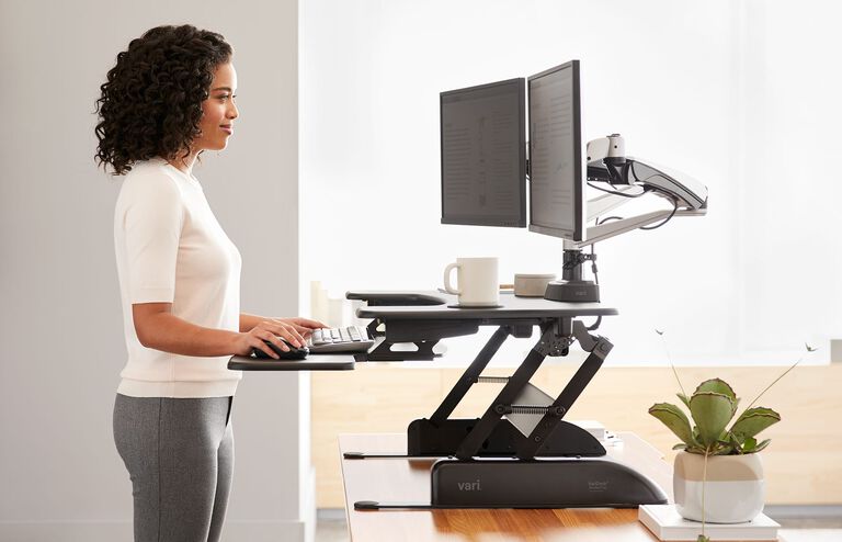 A sideview of an individual is standing at a raised converter ontop of a desk working in an office setting