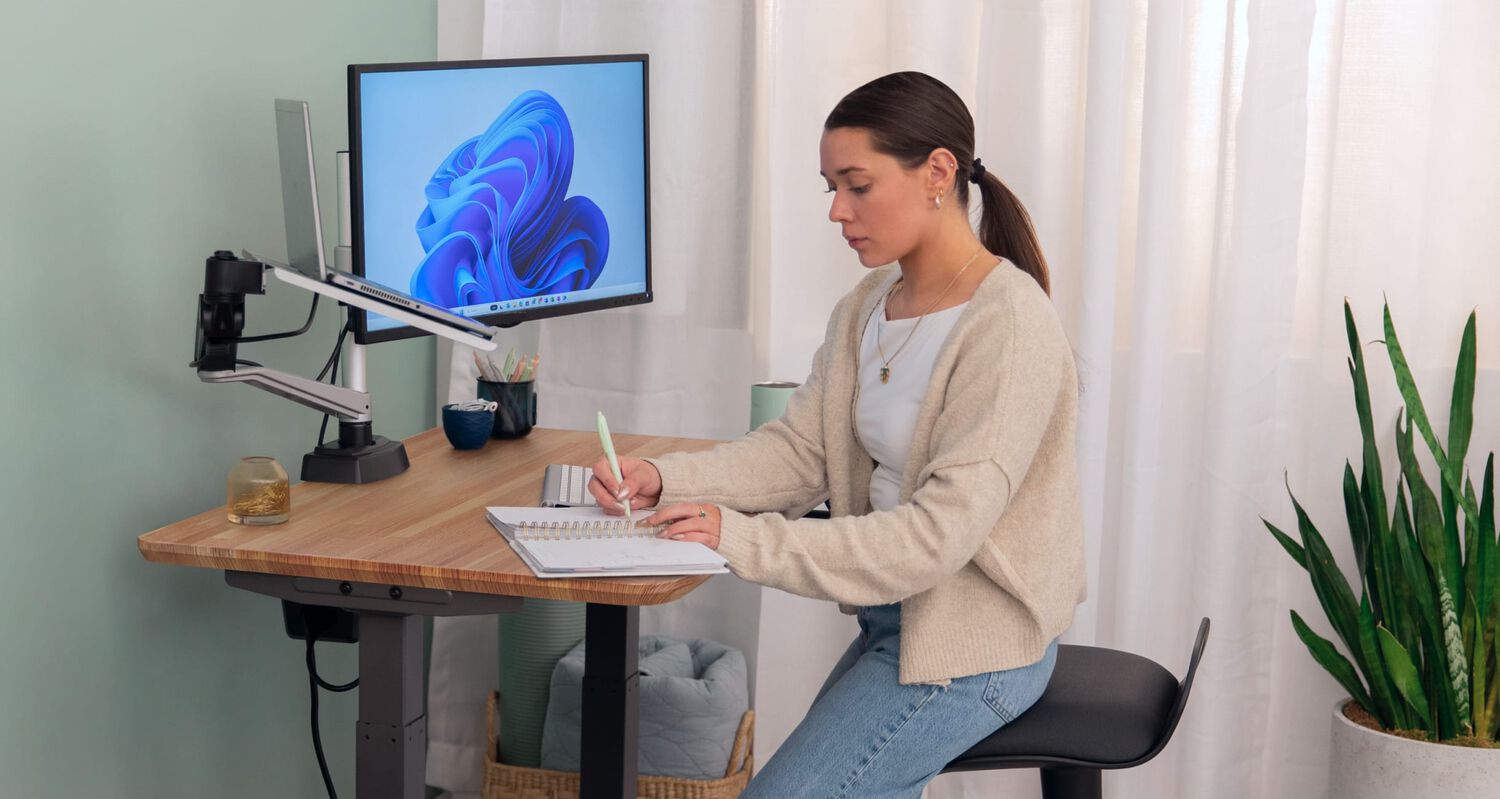 a woman sitting and writing in a notebook at an electric standing desk