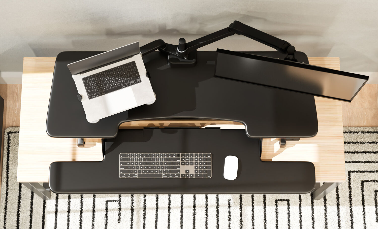 Top-down view of a black electric standing desk converter with a laptop tray, monitor arm, keyboard, and mouse, on a wood desk with a striped rug below.