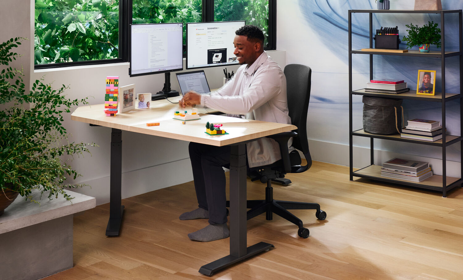 Man sitting on the Vari TempurPedic Chair at a height-adjustable desk in a home office, smiling while building with colorful blocks. He's barefoot in a black ergonomic chair, with dual monitors, personal items, and shelves with books and decor in the background.