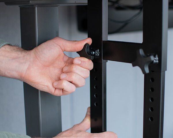 Close-up of hands adjusting a black metal desk mount by tightening a knob on a height-adjustable bracket with multiple holes.