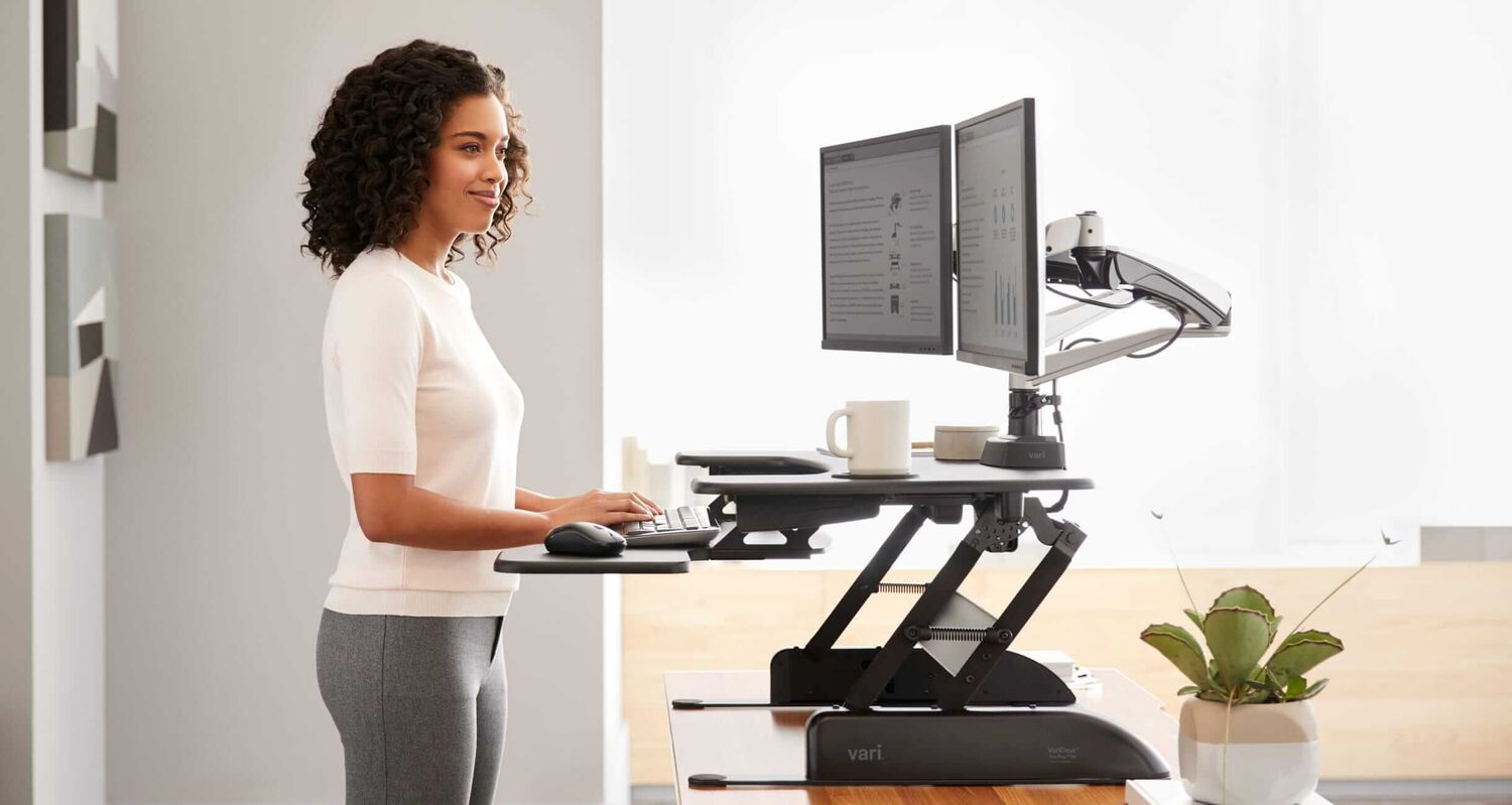 professional woman using a varidesk standing desk converter with dual monitor arm displays and a task chair in an office setting