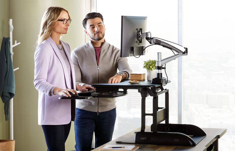 Two people standing at desk converter raised to max height working and looking at the single monitor mounted to the converter