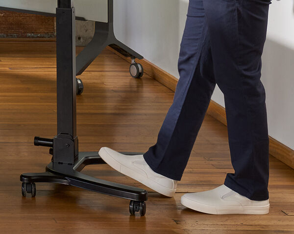 Close-up of a person in white sneakers rolling the mobile desk's four-star caster base across a wood floor, illustrating its mobility.