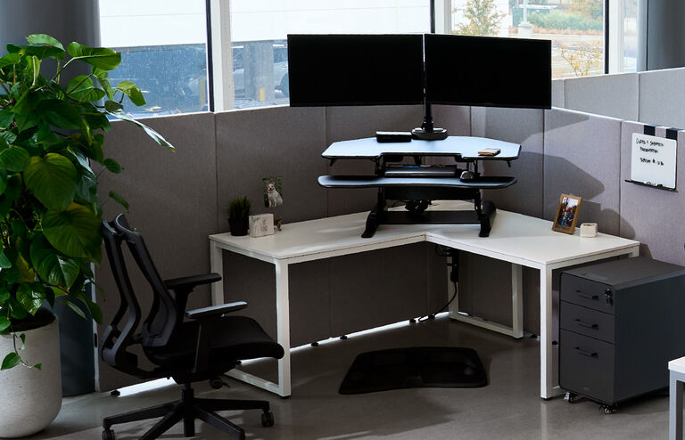 VariDesk Cube Corner in a modern office cubicle setup featuring a white l-shaped table with a black standing desk converter holding dual monitors. A black ergonomic office chair is positioned in front of the desk, and a black anti-fatigue mat is on the floor. The workspace is enclosed by gray fabric partition cubicle walls, with a small whiteboard displaying notes. Personal touches include a potted plant, a framed photo, and a small decorative sign. A slate rolling file cabinet is placed beside the desk for storage. A large green plant sits in a white pot nearby, adding a touch of nature to the workspace. Natural light streams in through large windows, providing a bright and comfortable environment.