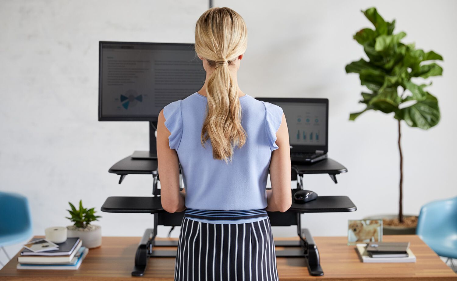 professional woman using a varidesk standing desk converter with dual monitor arm displays and a task chair in an office setting