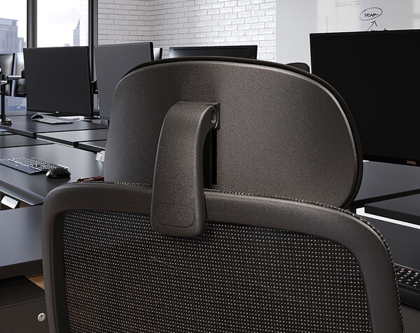 Close-up rear view of a black headrest and its adjustable mounting bracket attached to a mesh office chair, in an open office with a monitor and whiteboard.