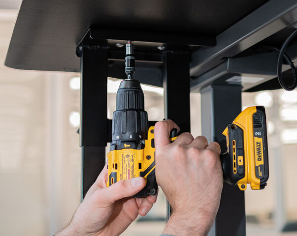 Person using a power drill to secure a screw under a black metal desk frame, focusing on the hands and tool in action.