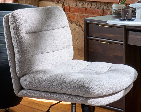 Close-up angled view of the light gray boucle armless office chair, showing the channel-tufted backrest and cushioned seat, in front of a desk with an exposed brick wall in the background.
