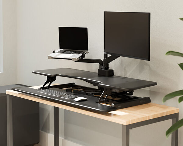 Black electric standing desk converter raised on a wood desk, holding a laptop and monitor, with a keyboard and mouse on the lower deck near a window and plant.