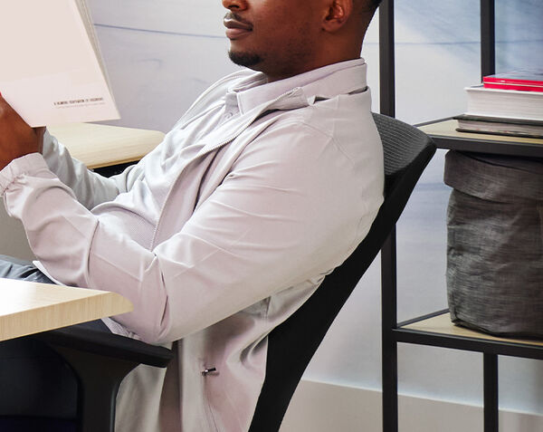 Man sitting in a modern Vari TempurPedic ergonomic office chair with a mesh back, reclining comfortably while reading a book at a desk in a contemporary office space with shelving and books in the background.