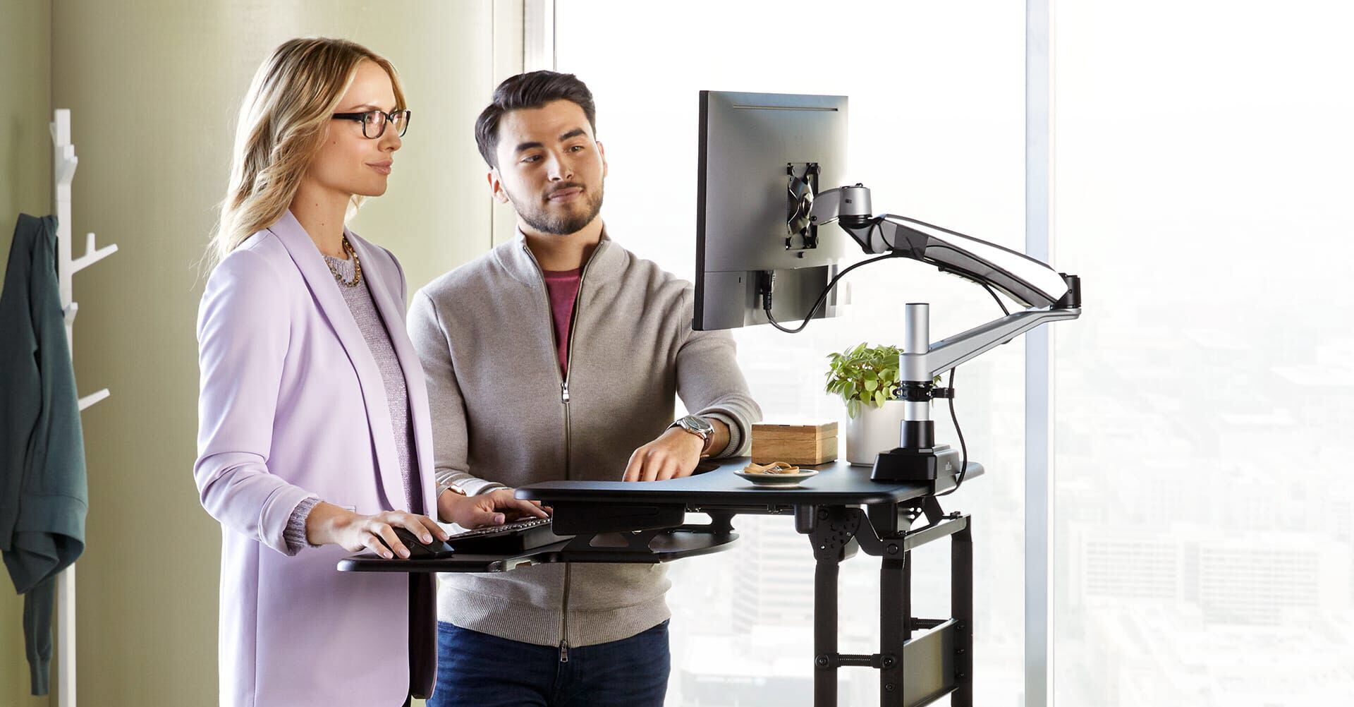 Two people standing at desk converter raised to max height working and looking at the single monitor mounted to the converter