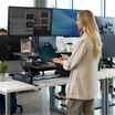 A professional office environment featuring a woman with long blonde hair using a black standing desk converter on a white desk. She is typing on a laptop, with two large monitors mounted above her workspace. The desk setup includes a framed photo, a small potted plant, and an ergonomic keyboard tray. In the background, other employees are working at similar standing desk setups, with modern office furnishings, large windows, and natural lighting creating a bright and collaborative atmosphere.
