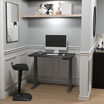 Black electric standing desk lowered in a stylish home office nook with a monitor, keyboard, and ergonomic stool.