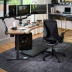 Modern home office setup featuring a black ergonomic Vari Tempur-Pedic chair at a curved wood desk with triple monitor arms, editing equipment, camera gear, and a gray area rug on hardwood flooring.