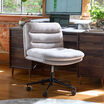 Light gray boucle armless office chair with a channel-tufted back and black five-star caster base, positioned in front of a dark wood desk with a monitor, laptop, and desktop converter, in a loft-style office with brick walls and large windows.