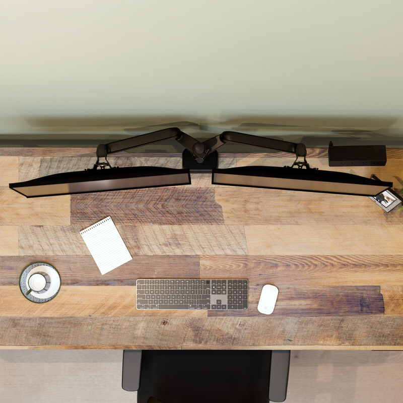 Top-down view of two monitors mounted on a dual monitor arm extending from a central pole, alongside a keyboard, mouse, notepad, and teacup on a wood desk. image number null