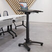Mobile adjustable-height laptop desk with an open laptop on the black tabletop, positioned in an open office space near a white conference table and mesh-back chairs, set against a wood slat accent wall.