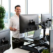 A professional office environment featuring a man standing up using a black standing desk converter on a white desk. He is talking on a video call, with two large monitors mounted above his workspace. 