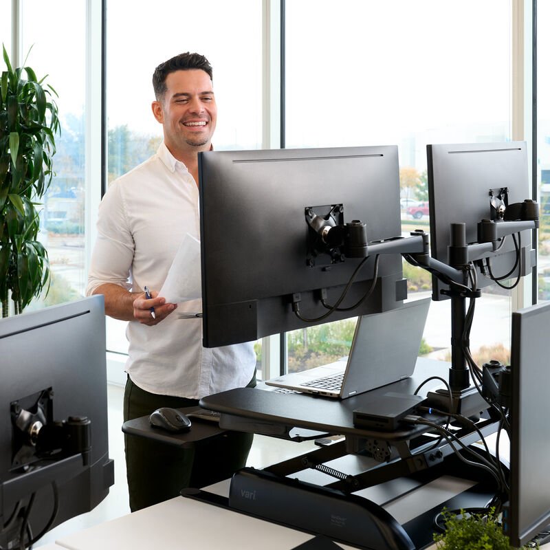 A professional office environment featuring a man standing up using a black standing desk converter on a white desk. He is talking on a video call, with two large monitors mounted above his workspace.  image number null