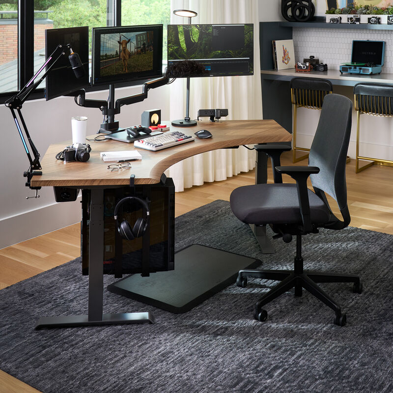 Modern home office setup featuring a dark gray ergonomic Tempur-Pedic chair at a curved wood desk with triple monitor arms, editing equipment, camera gear, and a gray area rug on hardwood flooring. image number null