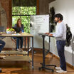 Man in a white sweater standing at the mobile laptop desk with his hand on the laptop trackpad, speaking to a woman with glasses seated at a nearby table, with a whiteboard labeled "Warehouse" visible behind him in a brick-walled office space.