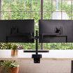 Dual monitor arm mounted to a desk holding two black monitors side by side, with a desk clamp base, in a home office with a picture frame, keyboard, and mouse.