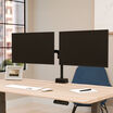 Rear view of a black dual monitor arm holding two monitors, showing the crossbar, pole, and desk clamp mount, in front of a window with greenery.