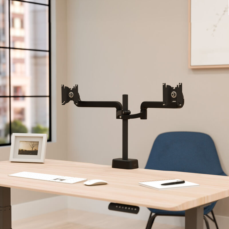 Front view of a black dual monitor arm without monitors attached, showing the two swing arms, center pole, and desk clamp base, on a desk with a keyboard and picture frame. image number null