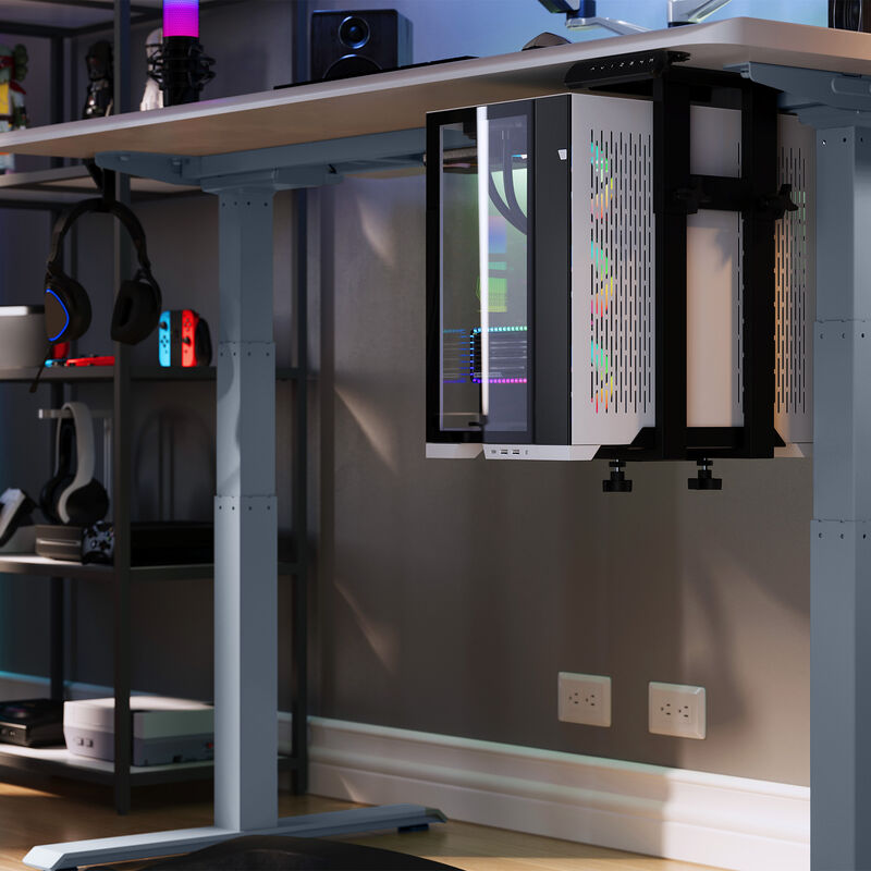 A gaming PC mounted under a sit-stand desk using a black Vari desk mount; RGB lighting is visible inside the PC case, with a tech setup and gaming accessories in the background. image number null