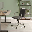 Modern office space with a sleek black ergonomic chair on casters, light wood flooring, large industrial windows, and minimalist furniture against an exposed brick wall backdrop.