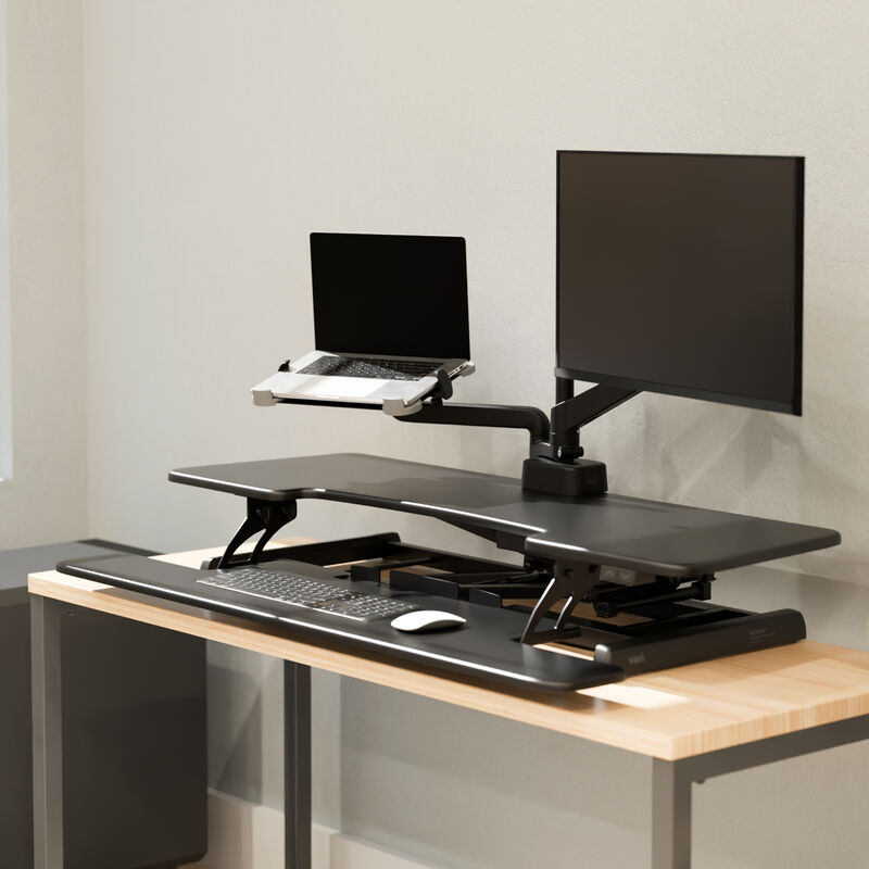 Black electric standing desk converter raised on a desk, with a laptop tray and monitor arm holding a laptop and monitor, plus a keyboard and mouse on the lower deck. image number null